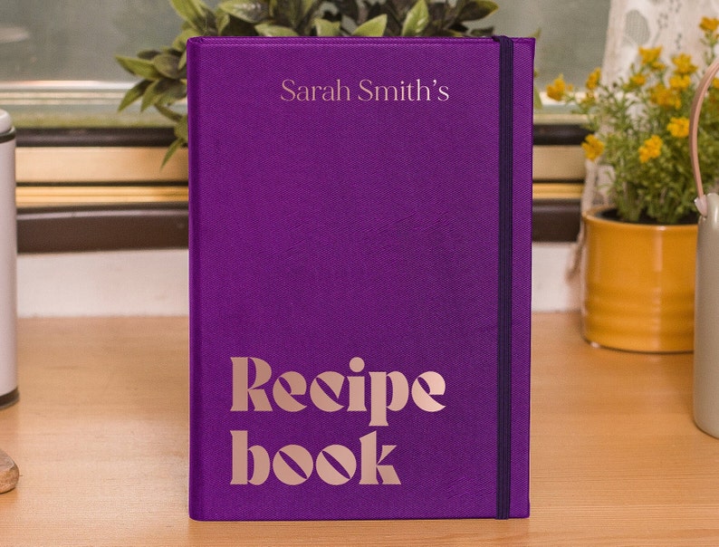 Puede incluir: Libro de recetas morado con el texto "Sarah Smith's Recipe Book" en letras doradas.
