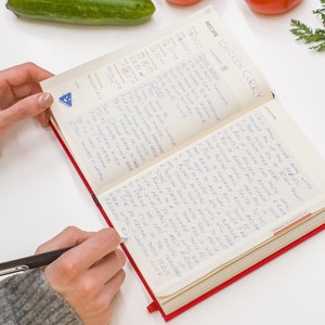 Puede incluir: Un libro de recetas abierto con instrucciones escritas a mano para hacer pollo al curry. El libro tiene una cubierta roja y est&aacute; abierto en una p&aacute;gina con el t&iacute;tulo "Pollo al curry" escrito en tinta azul.