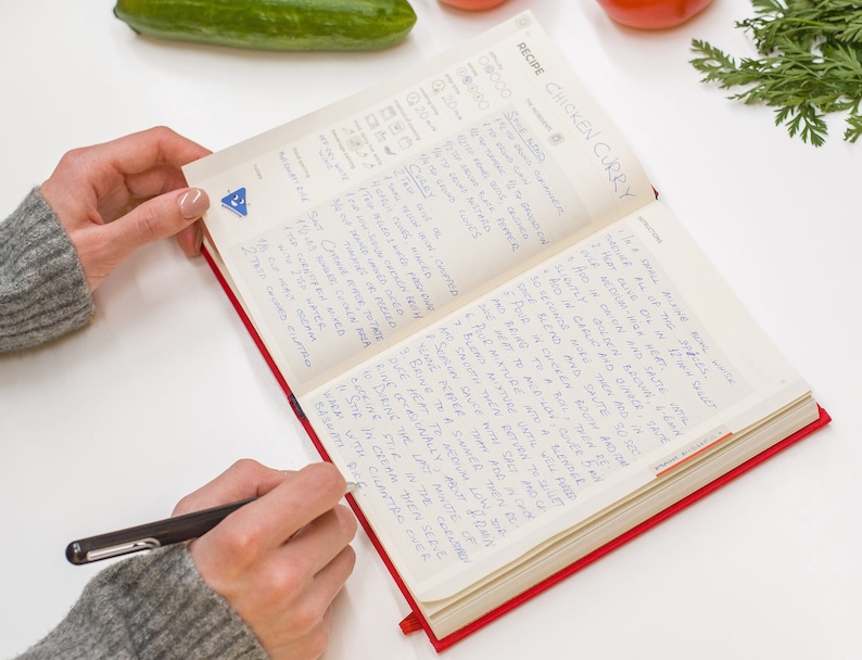 Puede incluir: Un libro de recetas abierto con instrucciones escritas a mano para hacer pollo al curry. El libro tiene una cubierta roja y est&aacute; abierto en una p&aacute;gina con el t&iacute;tulo "Pollo al curry" escrito en tinta azul.