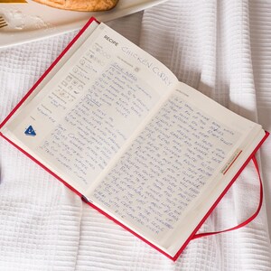Puede incluir: Un libro de recetas rojo abierto con instrucciones escritas a mano para un curry de pollo. La receta incluye ingredientes como especias, leche de coco y crema. El libro est&aacute; sobre un mantel de lino blanco.