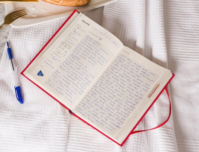 Puede incluir: Un libro de recetas rojo abierto con instrucciones escritas a mano para un curry de pollo. La receta incluye ingredientes como especias, leche de coco y crema. El libro est&aacute; sobre un mantel de lino blanco.