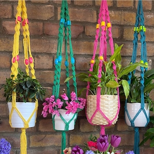 Puede incluir: Cuatro macramés para colgar plantas con cuentas de colores que cuelgan de una pared de ladrillo. Los macramés son de color amarillo, verde, rosa y azul. Cada macramé tiene una planta en una maceta.