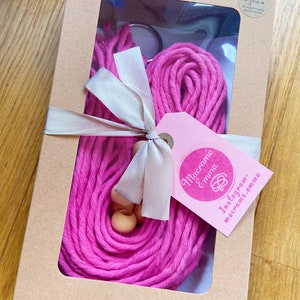 May include: A pink macrame cord kit in a brown box with a white ribbon and wooden beads. The box has a clear window showing the cord and beads. The box also has a tag that says "Macrame Emma" and "Instagram - macrame.emma".
