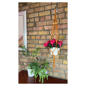 May include: A handcrafted macrame plant hanger in a warm brown color, adorned with wooden beads, suspends a white ceramic pot filled with vibrant pink flowers. The plant hanger is suspended from a metal ring, adding a touch of bohemian style to the space.