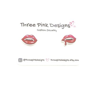 May include: A pair of red and pink lip-shaped earrings with fangs, one with a drop of blood. The earrings are on a white card with the text "Three Pink Designs custom jewelry" and social media handles.