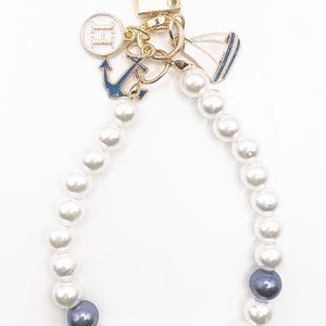 May include: A white pearl keychain with a gold anchor charm, a gold sailboat charm, and a gold charm with the letter "H" in a circle. The keychain has two dark gray pearls.