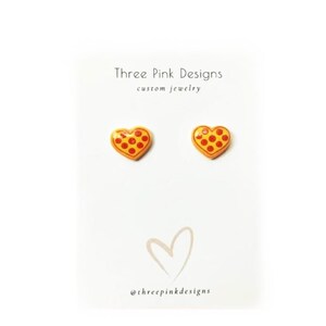 May include: Heart-shaped pizza earrings with red pepperoni on a yellow background. The earrings are displayed on a white card with the text "Three Pink Designs custom jewelry" and a heart graphic. The @threepinkdesigns Instagram handle is also visible.
