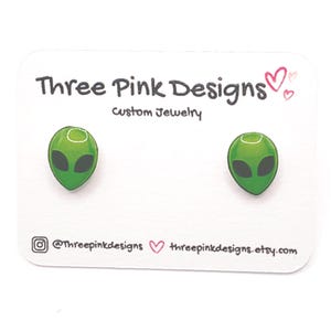 May include: A pair of alien-shaped stud earrings. Each earring features a green alien head with black eyes. The earrings are displayed on a white card with the text "Three Pink Designs Custom Jewelry". The card also includes social media handles.
