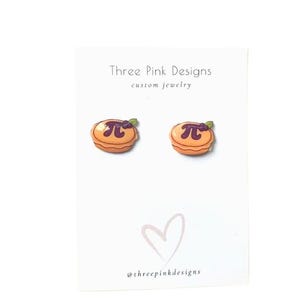 May include: A pair of novelty earrings shaped like miniature pies, each with a purple berry topping and a pi symbol. The earrings are on a white card with the text "Three Pink Designs custom jewelry" and a heart graphic.