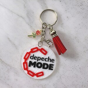 May include: A white circular keychain with the text "Depeche Mode" in black lettering and a red chain design. The keychain has a silver key ring with a red tassel and a small gold rose charm.
