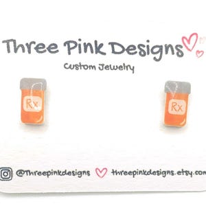 May include: A pair of orange and gray prescription bottle stud earrings. The earrings are on a white card with the text "Three Pink Designs Custom Jewelry". The card also includes social media information.