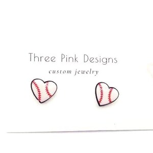 May include: Heart-shaped earrings with baseball stitching detail, set against a white backdrop. The text reads "Three Pink Designs custom jewelry". These earrings are a unique accessory.