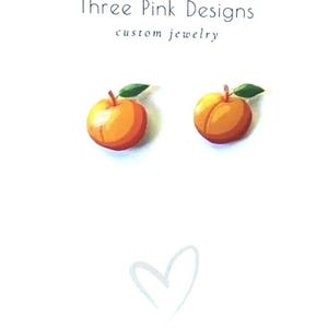 May include: Two peach-shaped earrings with green leaves and red stems are displayed on a white background. The text "Three Pink Designs custom jewelry" is at the top, and the Instagram handle is at the bottom.