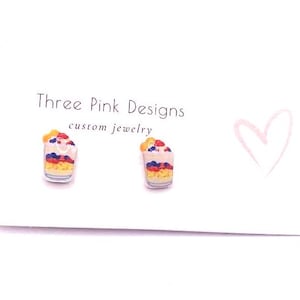 May include: A pair of stud earrings shaped like miniature parfaits, featuring layers of yellow, pink, and blue with red and yellow toppings. The earrings are displayed on a white card with the text "Three Pink Designs custom jewelry" and a pink heart.