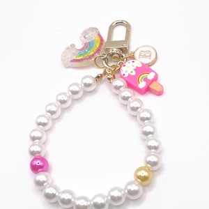 May include: A white pearl keychain with a pink pearl, a gold pearl, a rainbow charm, and a pink popsicle charm with a rainbow and clouds. The keychain has a gold ring and a gold letter "B" charm.