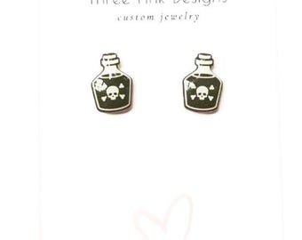 May include: A pair of black and white poison bottle earrings with a skull and crossbones design. The earrings are made by Three Pink Designs, a custom jewelry company.