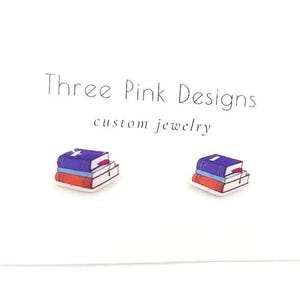 May include: A pair of stud earrings featuring a stack of three books with a purple, blue, and red cover. The earrings are on a white card with the text "Three Pink Designs custom jewelry".