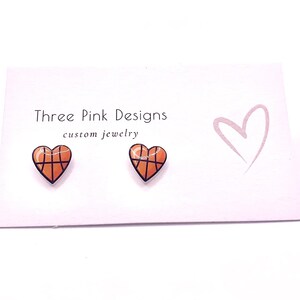 May include: A pair of heart-shaped earrings with a basketball design. The earrings are orange and brown with black lines. The earrings are on a white card with the text "Three Pink Designs custom jewelry".