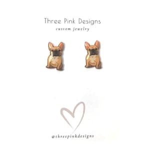 May include: A pair of stud earrings featuring a cartoon image of a French bulldog. The bulldog is light brown with black ears and nose. The earrings are made of resin and are approximately 1/2 inch in diameter.