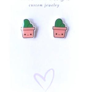 Könnte beinhalten: Ein Paar Kaktus-Ohrringe. Jeder Ohrring zeigt einen grünen Kaktus in einem rosa Topf mit einem lächelnden Gesicht. Die Ohrringe sind auf einer weißen Karte mit dem Text "Three Pink Designs custom jewelry".