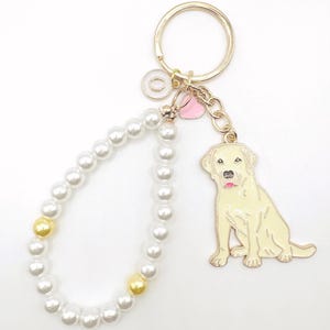 May include: A gold key chain with a white pearl bracelet, a pink heart charm, and a yellow Labrador Retriever charm.