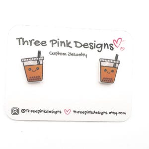 May include: A pair of stud earrings featuring a cartoon boba tea design. The earrings are on a white card with the text "Three Pink Designs Custom Jewelry". The earrings are brown and white with a smiling face.