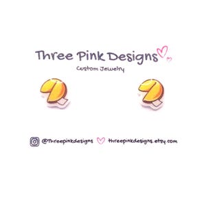May include: A pair of fortune cookie earrings on a white background. The earrings are yellow and white, with a cartoon-like design. The text "Three Pink Designs Custom Jewelry" is at the top, with social media handles at the bottom.