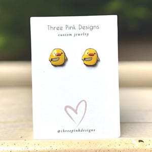 May include: A pair of yellow chick-shaped stud earrings with red accents, each holding a small knife. The earrings are displayed on a white card with the text "Three Pink Designs custom jewelry" and a heart graphic. The brand's Instagram handle is also visible.