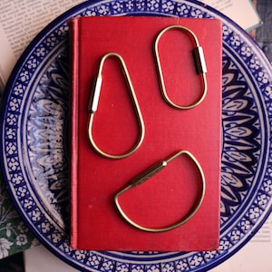 Könnte beinhalten: Drei Schlüsselanhänger aus Messing in verschiedenen Formen liegen auf einem roten Buch, das auf einem blau-weiß gemusterten Teller liegt. Die Schlüsselanhänger haben ein tropfenförmiges, ovales und halbkreisförmiges Design. Im Hintergrund befinden sich ein Vintage-Bügeleisen und ein offenes Buch.
