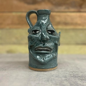 May include: A teal-colored ceramic jug with a face sculpted on the front. The jug has a handle and a spout. The face has a wide mouth, a furrowed brow, and a slightly tilted head. The jug is sitting on a light brown base.