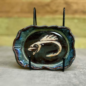 May include: A small, oval-shaped ceramic dish with a dark brown center and a teal and blue rim. The dish features a cream-colored, stylized sea serpent design. The dish is displayed on a black metal stand.