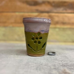 May include: A handmade ceramic votive candle holder. The top is a light purple color, and the bottom is green with a flower design cut out. A silver bottle cap sits next to the holder.