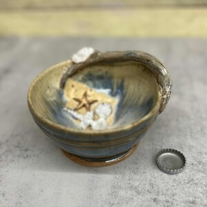May include: A handmade ceramic bowl with a coastal theme. The bowl is blue and tan with a starfish and seashell design. A bottle cap sits next to the bowl. The bowl has a textured rim.