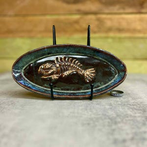 May include: An oval ceramic dish with a fish skeleton design. The dish has a dark brown center with a bronze-colored fish skeleton. The rim is a dark green with blue and bronze accents. The dish is displayed on a black metal stand.