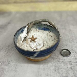 May include: A ceramic bowl with a blue rim and a handle. The interior features a sandy texture with a starfish and seashell design. A bottle cap sits beside the bowl. The bowl is a decorative item.