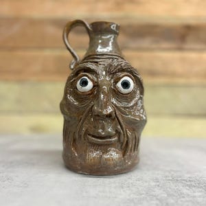May include: A brown ceramic jug with a handle, sculpted to resemble a weathered face with wide eyes. The jug has a glossy finish and detailed facial features, creating a rustic, folk art aesthetic.