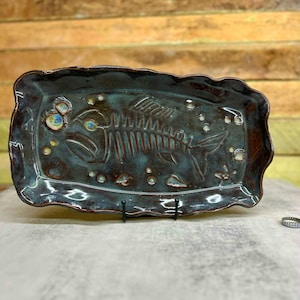 May include: Rectangular ceramic serving dish with a fish skeleton design. The dish has a dark teal glaze with brown edges and a wavy rim. Small, round, blue and white accents are scattered around the fish skeleton.