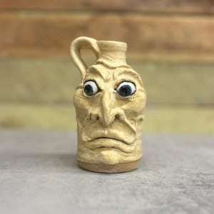 May include: A beige ceramic jug with a sculpted face. The jug features a handle and a detailed face with bulging eyes and a furrowed brow. The jug is approximately 5 inches tall.
