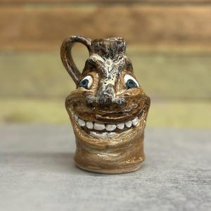 May include: A small, brown ceramic jug with a handle, featuring a smiling face design. The jug has a wide grin with white teeth, blue eyes, and a textured surface. The jug is a unique decorative item.