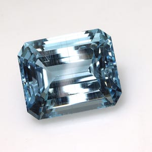 May include: A rectangular, faceted aquamarine gemstone with a light blue hue. The gemstone has an emerald cut with multiple facets that reflect light. The gem is set against a white background, highlighting its clarity and color.