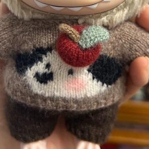 May include: A plush doll wearing a brown and white knit sweater with a red apple applique. The sweater has black spots and a pink nose. The doll's arms and legs are brown.