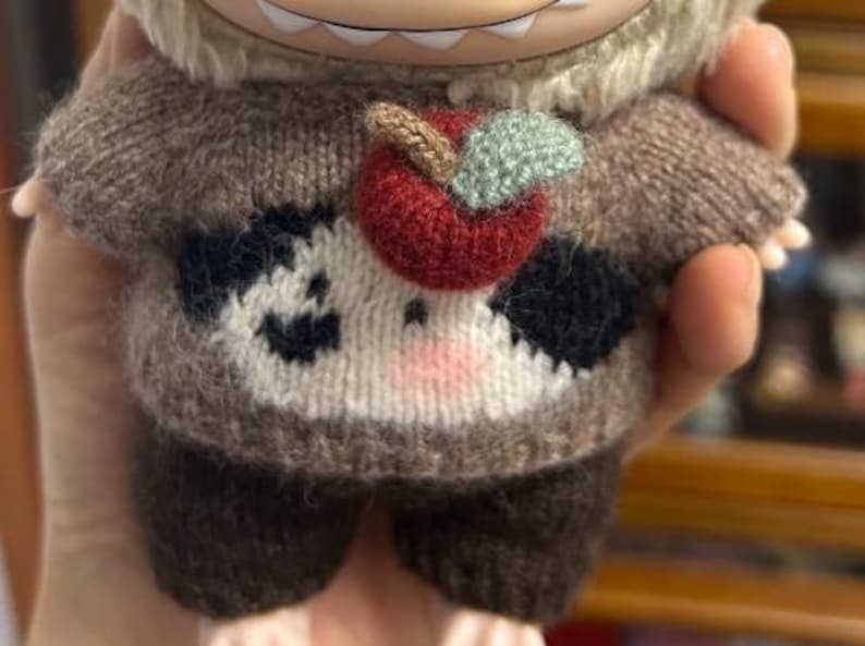 May include: A plush doll wearing a brown and white knit sweater with a red apple applique. The sweater has black spots and a pink nose. The doll's arms and legs are brown.