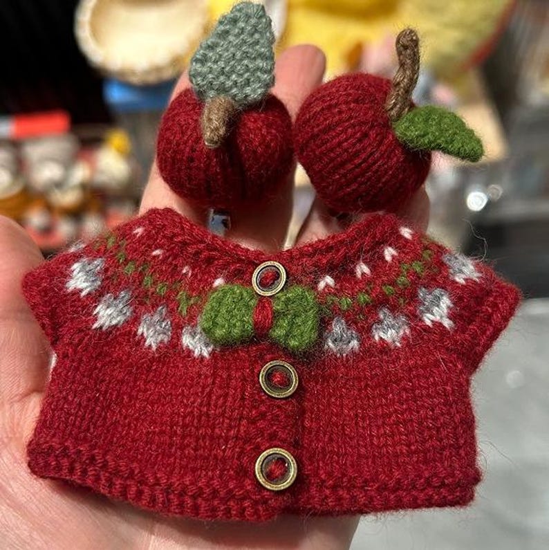 May include: A miniature red knitted sweater with a green bow and three gold buttons. Two knitted red apples with green leaves and brown stems are also visible. The sweater has a white and green pattern around the neckline.