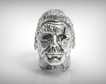May include: A silver-coloured ring featuring a detailed, realistic sculpture of a human face. The face has a cracked, weathered appearance, with a prominent scar across the forehead.