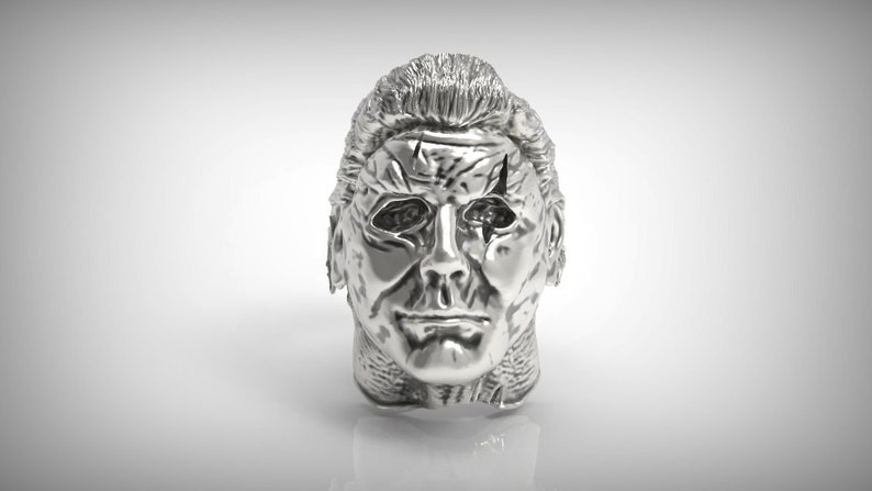 May include: A silver-colored ring with a detailed, realistic sculpture of a human face. The face has a cracked, weathered appearance, with a prominent scar across the forehead.