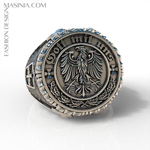 May include: A metallic ring featuring a central circular emblem with an eagle motif, oak leaves, and the text "Gott mit uns" engraved along the top edge. The sides of the ring display a cross symbol and intricate textured detailing in a dark finish.