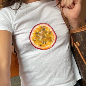 Pode incluir: Uma camiseta branca com um gráfico de uma fruta da paixão cortada ao meio. A fruta é amarela com sementes pretas e uma casca vermelha.