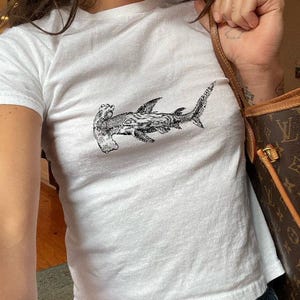 May include: White t-shirt with a black and white hammerhead shark illustration. The short-sleeved top is made of a soft fabric. The shark design is a detailed graphic, positioned centrally on the front of the shirt.