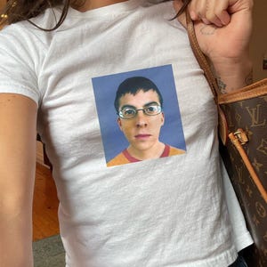 May include: A white t-shirt with a printed image of a person wearing glasses and a yellow and red shirt. The person is looking directly at the camera.
