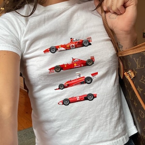 Könnte beinhalten: Weißes T-Shirt mit vier roten Formel-1-Rennwagen. Die Autos haben schwarze Räder und Sponsorenlogos. Das Shirt ist ein Crop-Top-Stil.